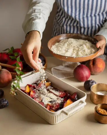 Person sprinkling a mixture over a dish of sliced fruits, with fresh peaches and plums on the table, demonstrating the use of guar gum in cooking.