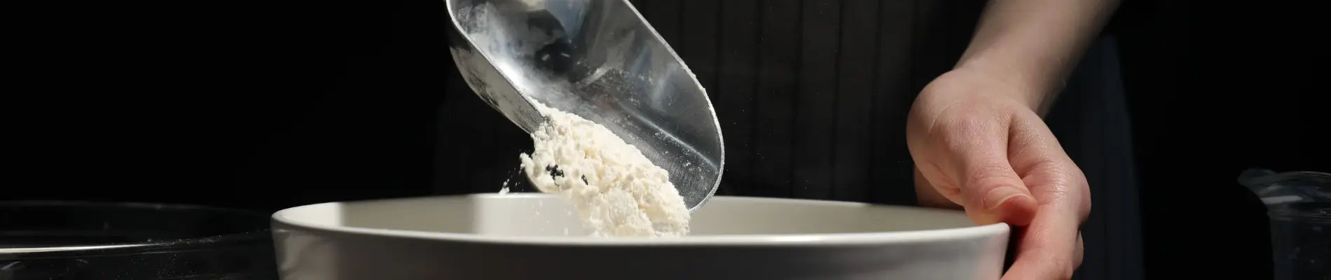 Person adding powdered ingredient into a bowl, illustrating the use of thickening or gelling agents in food processing.