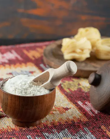 Wooden bowl filled with ULV guar gum powder and a small wooden scoop on colorful woven fabric, with dried food items in the background.